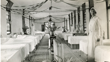 A black and white photograph featuring the internal of a ward in the Mental Hospital. Beds are arranged in two long lines, with a central aisle, and three nurses are in view.