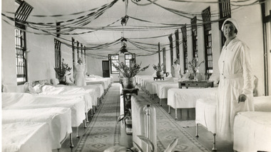 A black and white photograph featuring the internal of a ward in the Mental Hospital. Beds are arranged in two long lines, with a central aisle, and three nurses are in view.