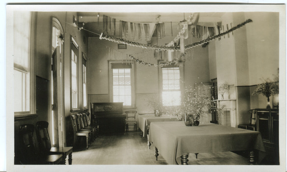 An empty dining room with two large tables down the centre of the room, with table cloths and a large vase of flowers on each. Chairs placed along the left wall with a piano in the far left corner. Paper decorations are hung across the room from the ceiling. Photograph has a white boarder.