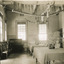 An empty dining room with two large tables down the centre of the room, with table cloths and a large vase of flowers on each. Chairs placed along the left wall with a piano in the far left corner. Paper decorations are hung across the room from the ceiling.