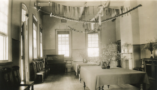 An empty dining room with two large tables down the centre of the room, with table cloths and a large vase of flowers on each. Chairs placed along the left wall with a piano in the far left corner. Paper decorations are hung across the room from the ceiling.