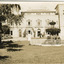 An outdoor photograph displaying the facade of the MayDay Hills Mental Hospital. The garden in front of the building shows a fenced fountain and large tree. The photograph has a white boarder with a strong crease in the bottom left corner.