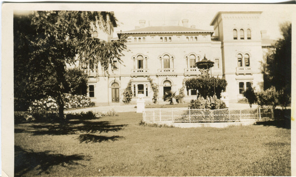 An outdoor photograph displaying the facade of the MayDay Hills Mental Hospital. The garden in front of the building shows a fenced fountain and large tree. The photograph has a white boarder with a strong crease in the bottom left corner.