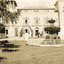 An outdoor photograph displaying the facade of the MayDay Hills Mental Hospital. The garden in front of the building shows a fenced fountain and large tree.