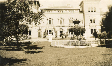 An outdoor photograph displaying the facade of the MayDay Hills Mental Hospital. The garden in front of the building shows a fenced fountain and large tree.