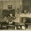 A postcard displaying an interior, featuring a large oval table with a vase of flowers in the centre. The table is flanked by two armchairs  and a fireplace in between and a large mirror above. The room also has two full bookcases off to the left. The postcard as a thin white boarder. 