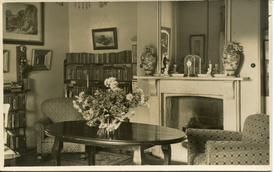 A postcard displaying an interior, featuring a large oval table with a vase of flowers in the centre. The table is flanked by two armchairs  and a fireplace in between and a large mirror above. The room also has two full bookcases off to the left. The postcard as a thin white boarder. 
