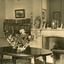 A postcard displaying an interior, featuring a large oval table with a vase of flowers in the centre. The table is flanked by two armchairs  and a fireplace in between and a large mirror above. The room also has two full bookcases off to the left. 