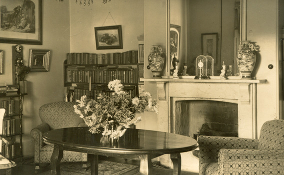 A postcard displaying an interior, featuring a large oval table with a vase of flowers in the centre. The table is flanked by two armchairs  and a fireplace in between and a large mirror above. The room also has two full bookcases off to the left. 