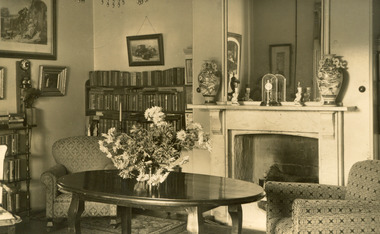 A postcard displaying an interior, featuring a large oval table with a vase of flowers in the centre. The table is flanked by two armchairs  and a fireplace in between and a large mirror above. The room also has two full bookcases off to the left. 