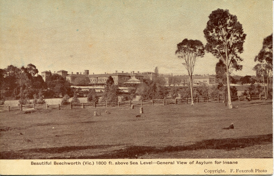A sepia toned image of a landscape with trees, farmland, and a wooden fence in the foreground, and the May Day Hills Mental Hospital building in the background, partly obscured by trees. A boarder on bottom edge with text saying "Beautiful Beechworth (Vic.) 1800 ft. above Sea Level - General View of Asylum for Insane" with "Copyright. F. Foxcroft Photo" underneath