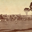 A sepia toned image of a landscape with trees, farmland, and a wooden fence in the foreground, and the May Day Hills Mental Hospital building in the background, partly obscured by trees.