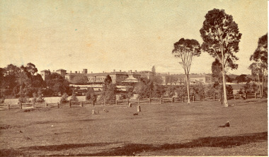 A sepia toned image of a landscape with trees, farmland, and a wooden fence in the foreground, and the May Day Hills Mental Hospital building in the background, partly obscured by trees.