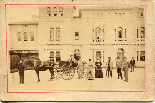 A child sitting in a horse drawn carriage surrounded by eight people in front of the Beechworth Mental Asylum's administration building 