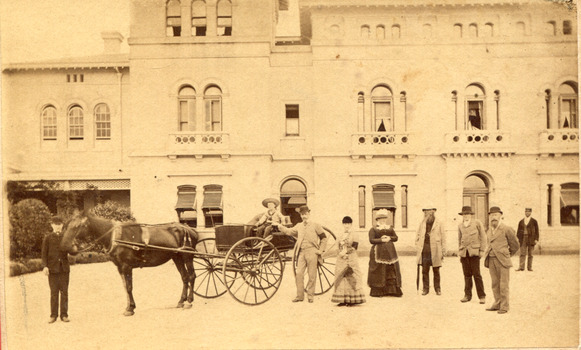 A child sitting in a horse drawn carriage surrounded by eight people in front of the Beechworth Mental Asylum's administration building 