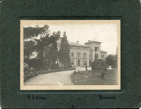 A pathway featuring manicured gardens on either side leading to the Beechworth Mental Asylum administration building. There are four males, three are sitting on the grass while one stands on one leg just behind them.