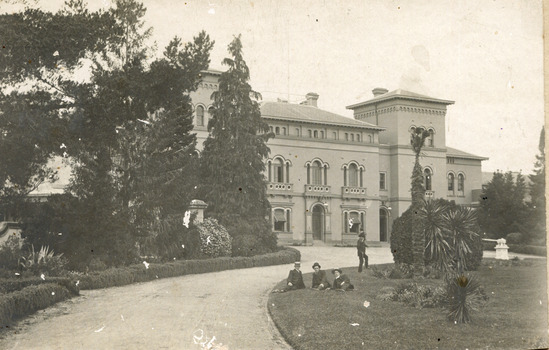 A pathway featuring manicured gardens on either side leading to the Beechworth Mental  Asylum administration building. There are four males, three are sitting on the grass while one stands on one leg just behind them.