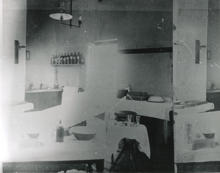 Grainy black and white photograph showing interior of surgery and consulting room in the Mental Hospital. Tables with medical instruments, bowls and jars are visible. Lefthand section of the photograph has been reproduced on the righthand side to show details which are lost due to overexposure. 