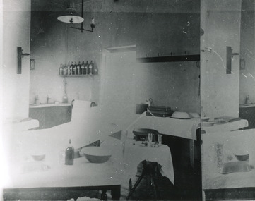 Grainy black and white photograph showing interior of surgery and consulting room in the Mental Hospital. Tables with medical instruments, bowls and jars are visible. Lefthand section of the photograph has been reproduced on the righthand side to show details which are lost due to overexposure. 