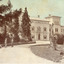 Colourised photograph postcard showing the front of the Mayday Hills asylum building with several large trees and grounds in front. In foreground of the image there is a person (possibly a boy) in a black suit and hat with their back to the camera leaning on their left elbow against a pedestal pillar looking back towards the building and at another two people who appear to be in conversation on the path leading to the asylum building.