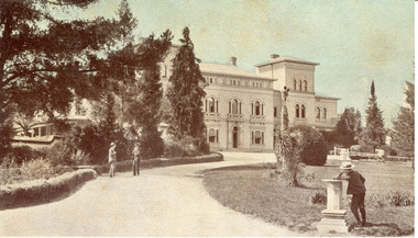 Colourised photograph postcard showing the front of the Mayday Hills asylum building with several large trees and grounds in front. In foreground of the image there is a person (possibly a boy) in a black suit and hat with their back to the camera leaning on their left elbow against a pedestal pillar looking back towards the building and at another two people who appear to be in conversation on the path leading to the asylum building.