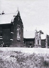 Black and white photograph of front view of Ovens Benevolent Home with one person sitting on a bench seat against the nearest part of the building and another person walking on the grass.
