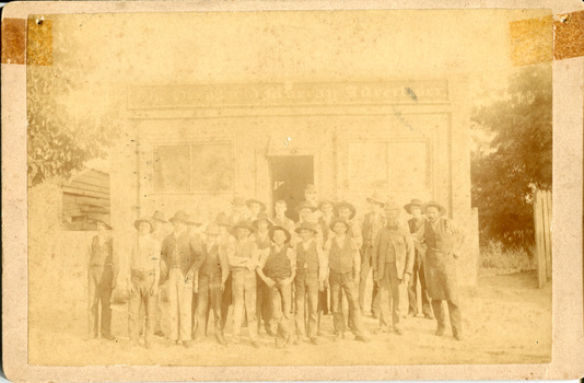 Group of workmen in hats standing in front of a building, the sign of which reads "The Owens & Murray Advertiser." 