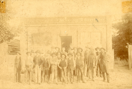 Group of workmen in hats standing in front of an old building, the sign of which reads "The Ovens and Murray Advertiser." 