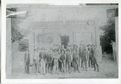 Group of workmen in hats standing in front of an old building, the sign of which reads "The Ovens and Murray Advertiser."