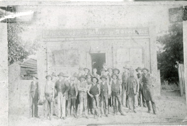 Group of workmen in hats standing in front of an old building, the sign of which reads "The Ovens and Murray Advertiser."