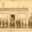 Group of men lined up horizontally in front of a building. The building sign reads "The Owens and Murray Advertiser." A bearded man is standing in the doorway. There is a fence and trees to teh right of the building.