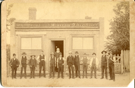 Group of men lined up horizontally in front of a building. The building sign reads "The Owens and Murray Advertiser." A bearded man is standing in the doorway. There is a fence and trees to teh right of the building.