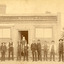Group of men lined up horizontally in front of a building. The building sign reads "The Owens and Murray Advertiser." A bearded man is standing in the doorway. There is a fence and trees to teh right of the building. 