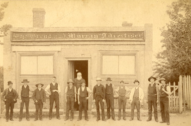 Group of men lined up horizontally in front of a building. The building sign reads "The Owens and Murray Advertiser." A bearded man is standing in the doorway. There is a fence and trees to teh right of the building. 
