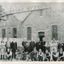 Group of young and old men in suits and aprons stand in front of a building, the sign of which reads "Ovens and Murray Advertiser Printing Office." There is a tree blocking the sign in the top-left quadrant  of the image, and a dog in the foreground.