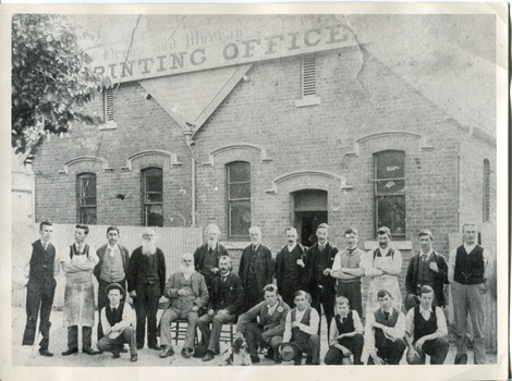 Group of young and old men in suits and aprons stand in front of a building, the sign of which reads "Ovens and Murray Advertiser Printing Office." There is a tree blocking the sign in the top-left quadrant  of the image, and a dog in the foreground.