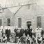 Group of young and old men in suits and aprons stand in front of a building, the sign of which reads "Ovens and Murray Advertiser Printing Office." There is a tree blocking the sign in the top-left quadrant  of the image, and a dog in the foreground. 
