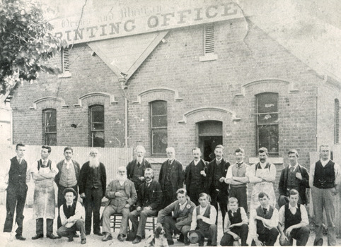 Group of young and old men in suits and aprons stand in front of a building, the sign of which reads "Ovens and Murray Advertiser Printing Office." There is a tree blocking the sign in the top-left quadrant  of the image, and a dog in the foreground. 