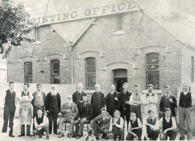 Group of young and old men in suits and aprons stand in front of a building, the sign of which reads "Ovens and Murray Advertiser Printing Office." There is a tree blocking the sign in the top-left quadrant  of the image, and a dog in the foreground. 