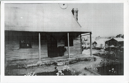 Small timber house with steep roof and flower beds in the front garden. Other houses or sheds can be seen in the background. 