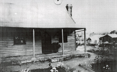 Small timber house with steep roof and flower beds in the front garden. Other houses or sheds can be seen in the background. 