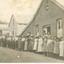 Sepia-toned photograph of a row of older and younger men and women standing in front of three buildings, two made of wood and one of rendered brick/stone and wood. In the doorway of the rendered building stands a man and woman. Above them, the building is marked as S.S.Jam.C.G.