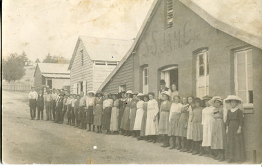Sepia-toned photograph of a row of older and younger men and women standing in front of three buildings, two made of wood and one of rendered brick/stone and wood. In the doorway of the rendered building stands a man and woman. Above them, the building is marked as S.S.Jam.C.G.