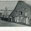 Uncropped black and white photograph on paper of a row of older and younger men and women standing in front of three buildings, two made of wood and one of rendered brick/stone and wood. In the doorway of the rendered building stands a man and woman. Above them, the building is marked as S.S.Jam.C.G.