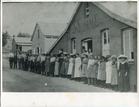 Uncropped black and white photograph on paper of a row of older and younger men and women standing in front of three buildings, two made of wood and one of rendered brick/stone and wood. In the doorway of the rendered building stands a man and woman. Above them, the building is marked as S.S.Jam.C.G.