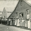 Black and white photograph of a row of older and younger men and women standing in front of three buildings, two made of wood and one of rendered brick/stone and wood. In the doorway of the rendered building stands a man and woman. Above them, the building is marked as S.S.Jam.C.G.