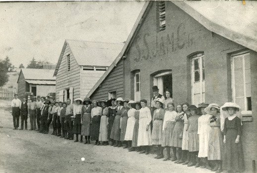 Black and white photograph of a row of older and younger men and women standing in front of three buildings, two made of wood and one of rendered brick/stone and wood. In the doorway of the rendered building stands a man and woman. Above them, the building is marked as S.S.Jam.C.G.