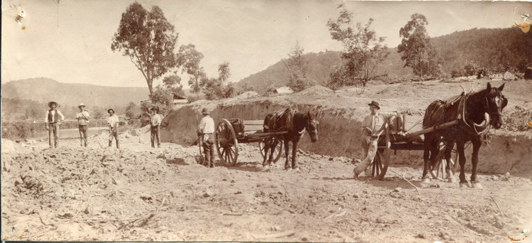 Six men and two horses with carts stand amidst a partially constructed road in the Australian countryside. Four men stand side by side approximately a metre apart. A fifth man stands beside one horse and cart with a shovel in his hand, facing the four. The last man stands in the foreground, leaning against the second horse and carriage.  