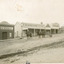 View of a group of businesses at Newtown, Beechworth. Businesses include Beechworth Foundry, R. McKenzie and Sons Grain Store, and McKenzie Family Store. In front of the grain store, a single horse drawn carriage, driven by one man, behind a second man on a horse. A dog stands beside the carriage.  