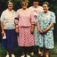 Four older women stand side by side smiling with trees and shrubbery behind them. 